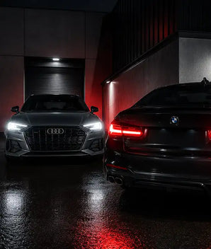 Eine elegante schwarze BMW-Limousine mit markanten roten Rücklichtern und einer glänzenden Oberfläche, die neben einem silbernen Audi-Sportwagen in einer schwach beleuchteten Garage steht.